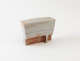 Rectangular ceramic sculpture with a pale, whitewashed surface flecked with rust colored specks, divided by a thin horizontal line above a rough, weathered red-brown base. 