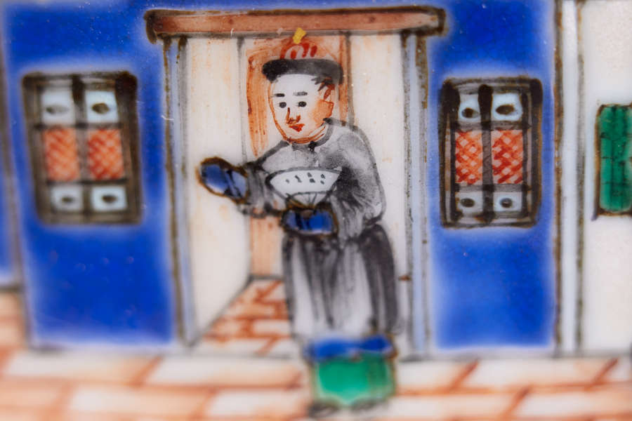 A close up view of a bowl with a painted figure, wearing a gray robe and holding a bowl in front of a doorway with blue walls and detailed windows.