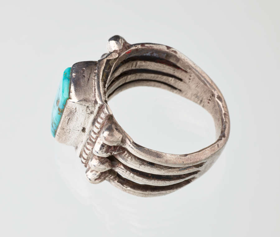 Angled side-view of a silver metal ring with a rectangular turquoise stone setting. Visible are three-dimensional embellishments of simple vegetal and rope motifs.