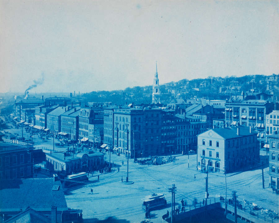 Blue-hued urban panorama, showcasing a street of buildings and a towering church steeple breaking the horizon. In the distance, an industrial chimney produces a large plume of dark smoke.