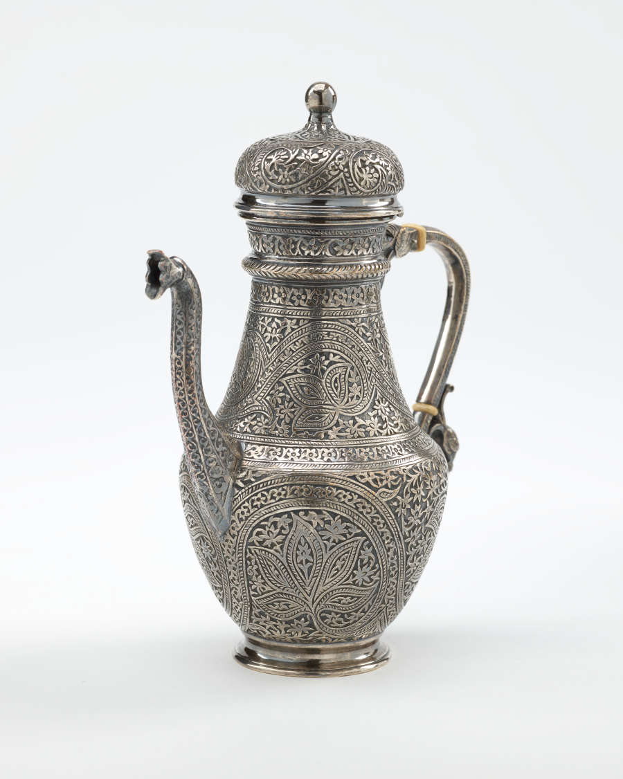 A silver coffee pot with detailed delicate decorations, handle with a lid hinged to it, and spout.