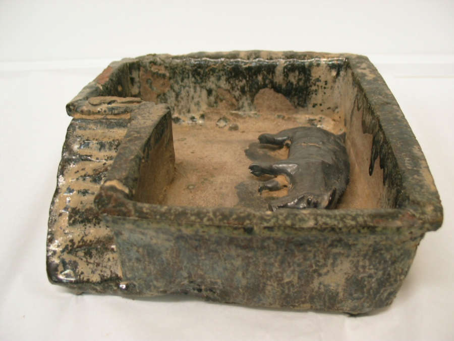 Ceramic sculpture showing a small pig laying on its side, enclosed by a rectangular structure which features arches and stairs. The sculpture is tan with a glossy muted green glaze. 