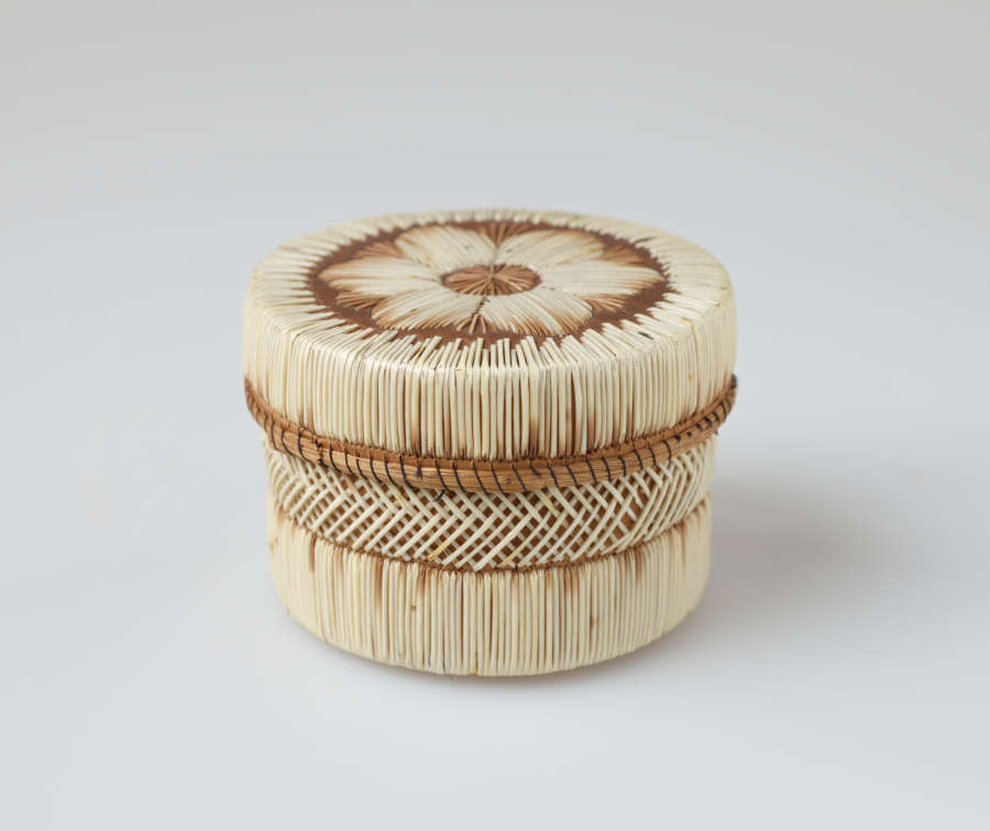 Cylindrical lidded leather box with cross-hatching and linear stripe patterns woven in white and brown straw. The brown rimmed lid has a central floral motif woven atop it.