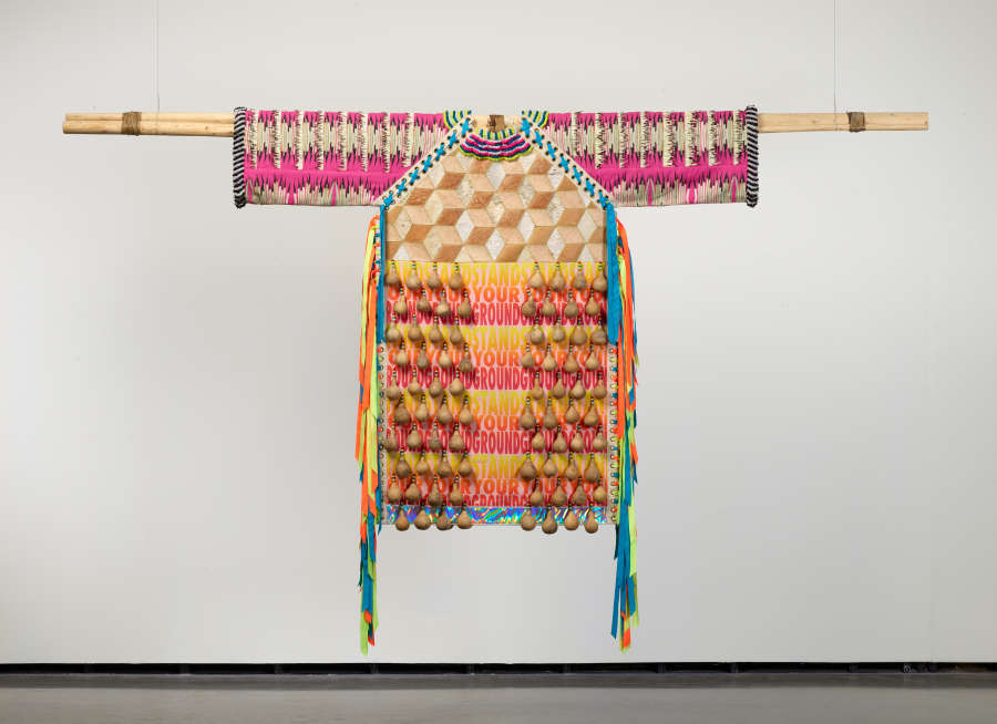 Multi-patterned, brightly colored, and tasseled shirt hanging on a wooden stick against a white background with machinery attached to its bottom. The floor is also shown.