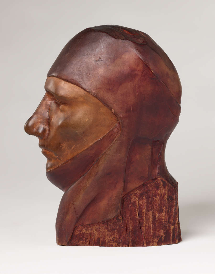 Profile view of a vibrant, carved wood sculpture depicting a serene face wearing a draped cloth around its head. It is smooth, besides a vertical crack alongside its nose.