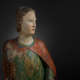 The face of a softly illuminated wooden statue, gaze turned slightly to the side with a serene expression, dressed in a faded red and green garment.