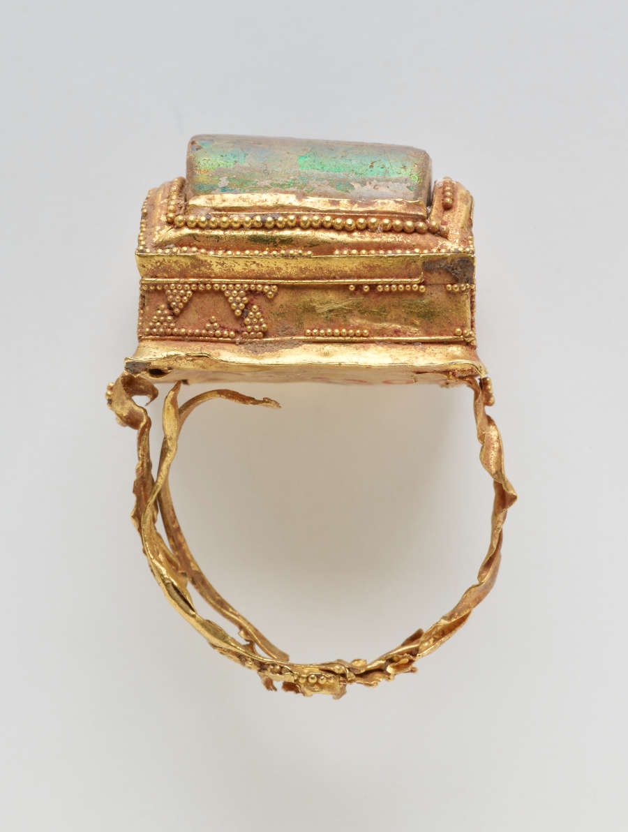 Ring, seen from the side. It seems old, fragile, and damaged. A green square stone is set in the top. The gold is bright and features delicate patterns.
