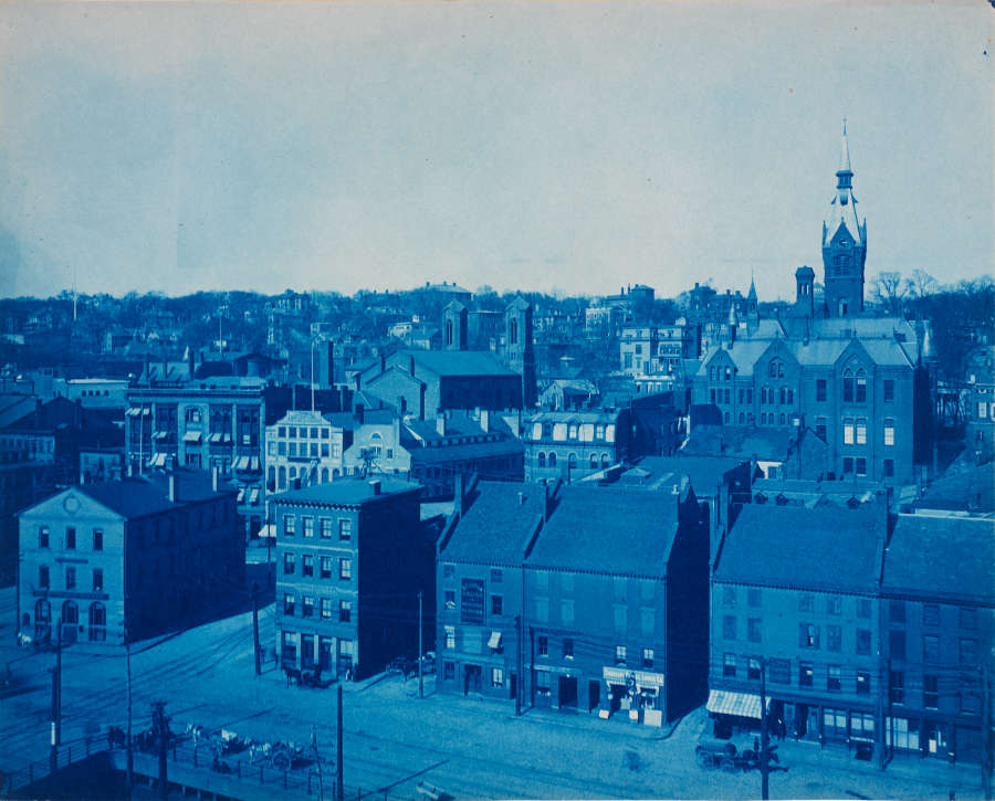 A blue tinted photograph captures a town center with historic brick buildings, behind which lies a hill. The streets are lined by storefronts, and the hill features a steepled building.
