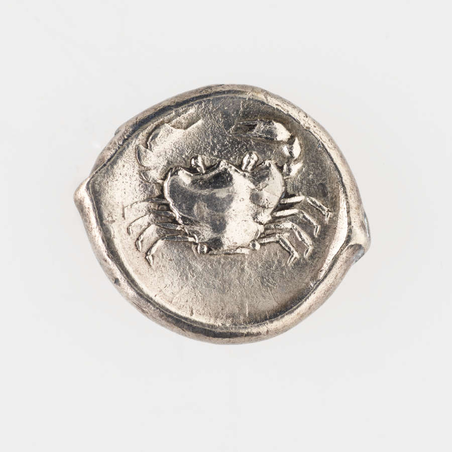 Silver coin with irregular borders. Embossed on one side is the top view of a crab. The edge of the coin is slightly raised.
