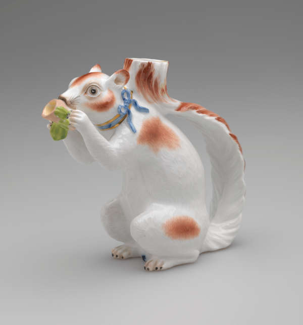 Vessel in the shape of a white squirrel with brown patches and a blue ribbon arounds its neck. It holds a brown nut with green leaves up to its face.