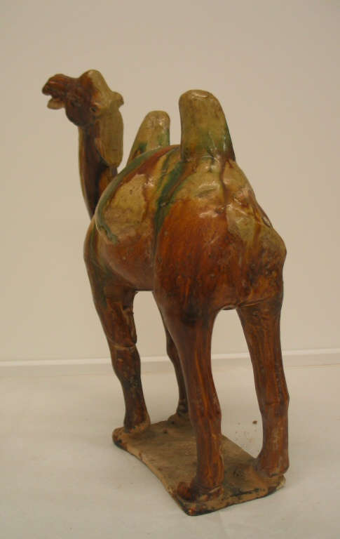 Angled back-view of a glossy green, tan, and brown figurine of a camel with two humps standing on a short attached rectangular platform.