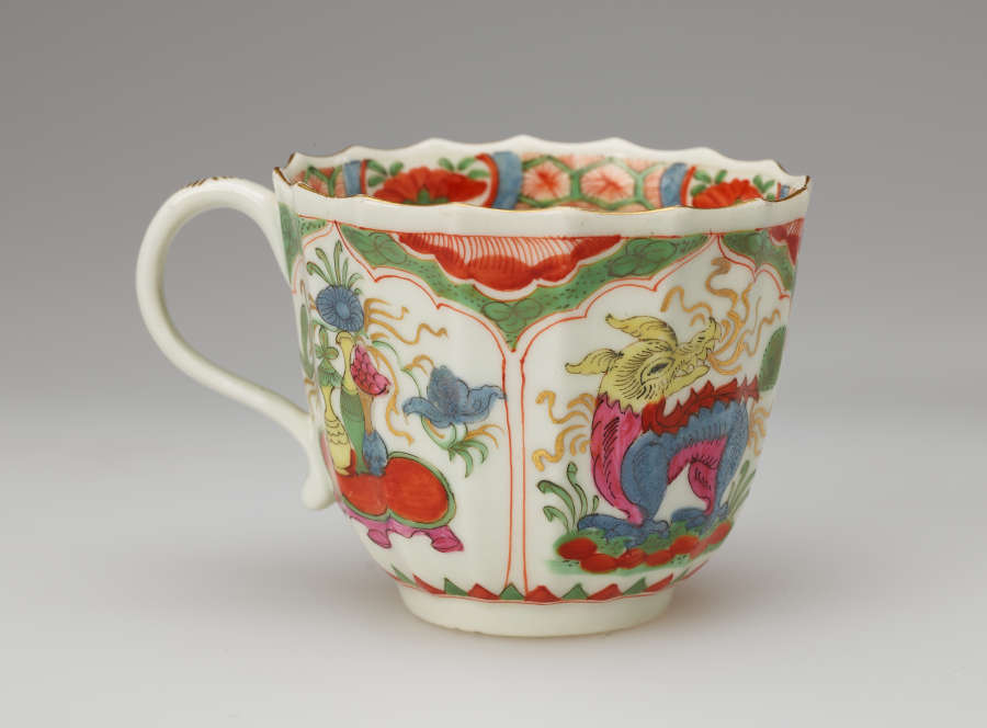 A white cup with a scalloped lip, green, red, blue, pink, yellow, gilded decorations, and two vignettes: an animal and the other a table with vessels on top of it.
