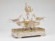 Angled side-view of elaborate silver double-oval centerpiece with swirling arms extending to two shallow side-dishes, with central fountain-like stem topped by classical figurines, all on a rectangular tray pedestal.  