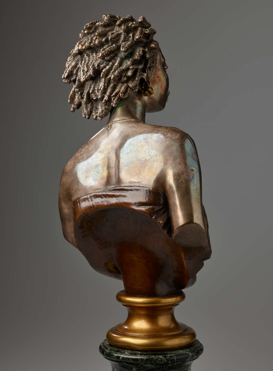 Back view of a bronze bust featuring hair in twists swept back and shoulder blades accentuated above a dark strapless top cut off diagonally and mounted on a gold base.