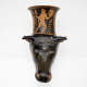 Black ceramic vessel shaped like a bull’s head with a flared cylindrical neck. The neck features an orange painted scene of a nude muscular figure holding a staff and shield. 