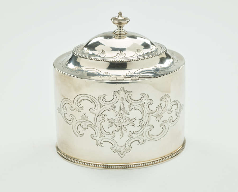 A silver tea caddy with a lid and sculptural finial. There are swirling engravings along the body and lid.