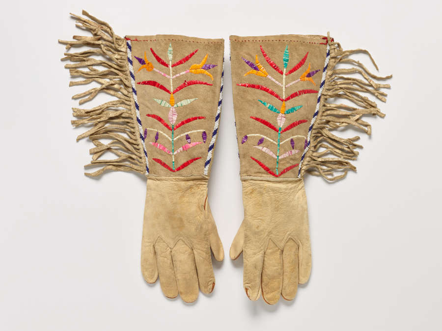 Pair of tan gloves laid flat, each with fringed and beaded, patterned edges, and colorful central symmetrical floral motifs embroidered onto the long cuffs.
