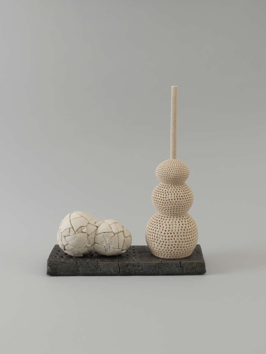 Two joined pale spheres with a cracked surface lie beside a vertical rod supporting a stack of three smooth, cream-colored spheres, all arranged on a black rectangular base.