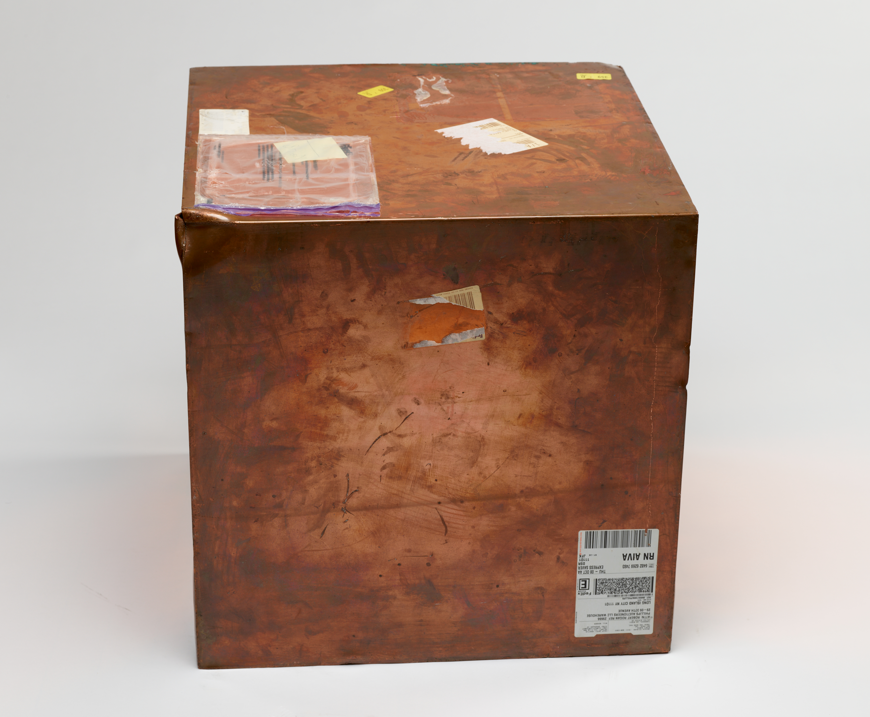 Image of Walead Beshty’s work: Copper box covered in various markings and shipping label stickers. The box is discolored in some areas.