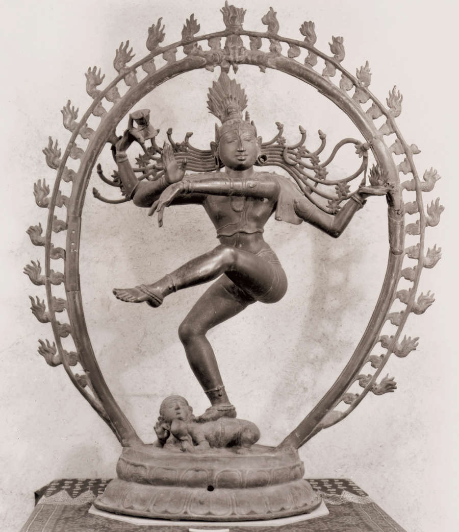 Dark bronze sculpture of a figure with one leg raised and the other bent, and one arm bent upwards and the other extending across its body, surrounded by a sculpted halo.