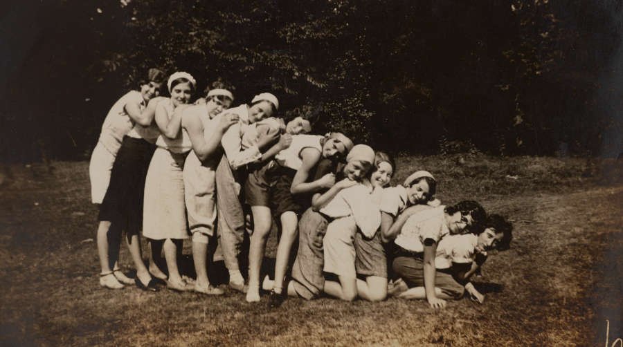 Affectionate snapshot of eleven smiling young women in a line, facing right. Each slumps onto the next. Those at left stand, those in the middle kneel; the last crouch underneath.