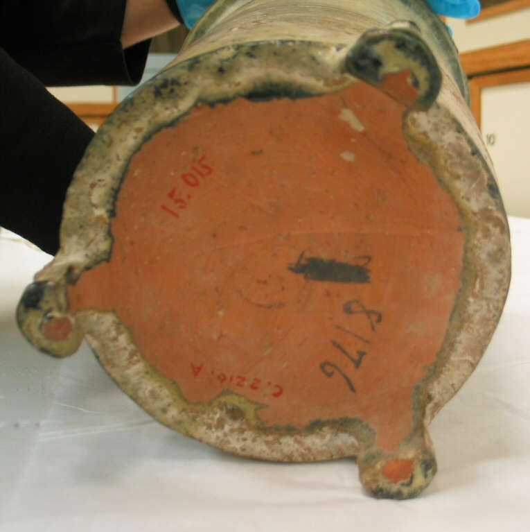 Round base of a terracotta urn in storage, with three semicircular feet emerging from its glazed edge. It bears red and black labels reading ‘8176’, ‘15.015’, and ‘C.2210:A’.