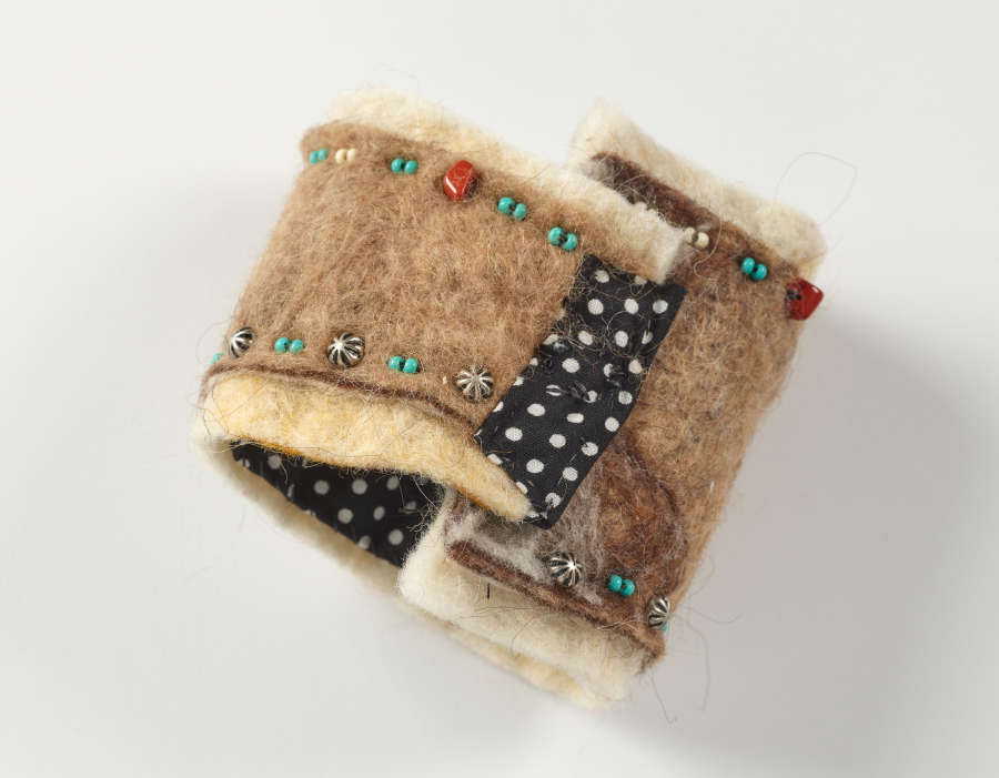 Top-view of a thick, clasped felted bracelet with silver embellishments and multicolored  beads. Black and white polka dot fabric lines the inside of the bracelet and clasps it shut. 