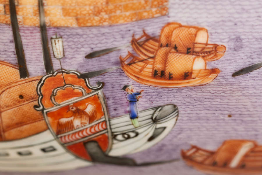 A close up of a painted ceramic bowl depicting a figure in blue standing on the deck of a decorated ship with woven coverings on purple water.