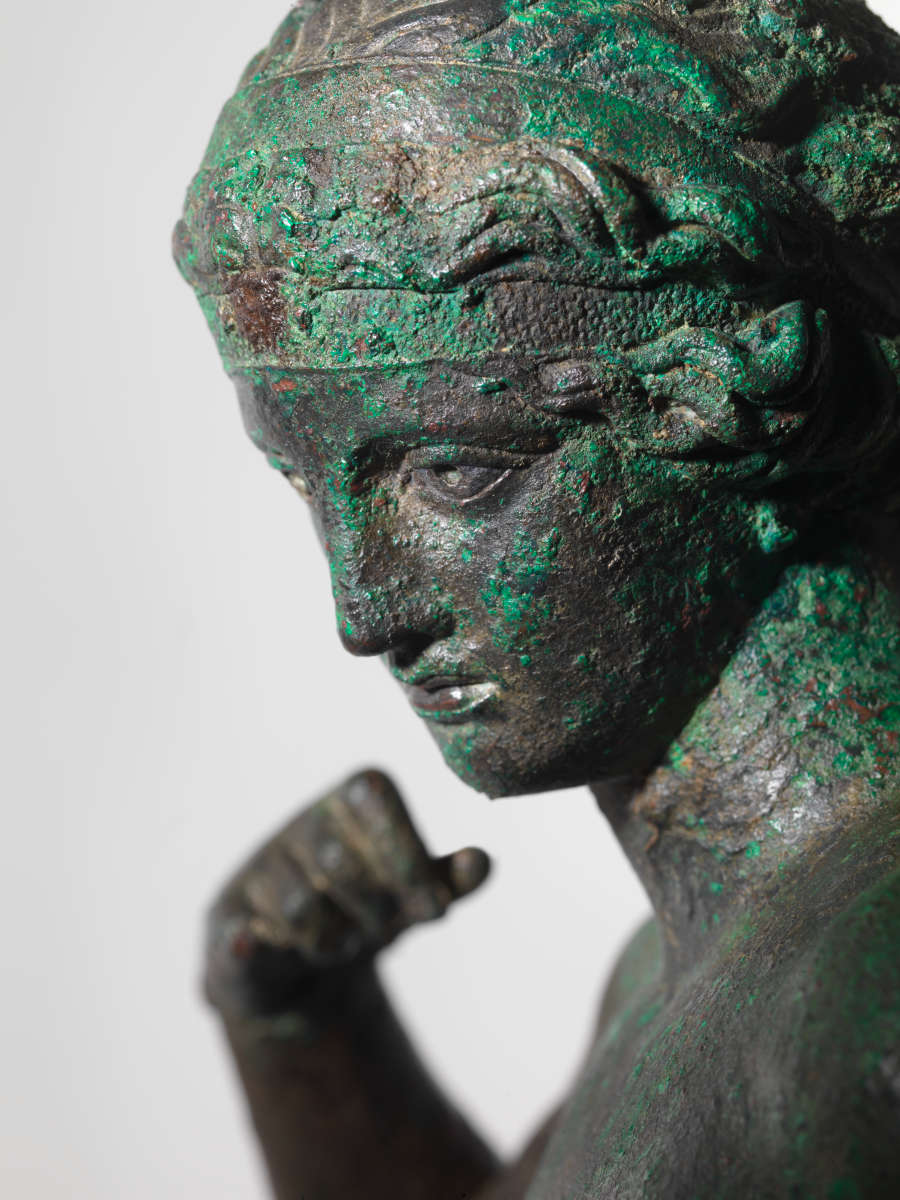 Detail view of a gray bronze statue of a crowned nude woman. Visible are the details of her face, hair and clenched fist curled towards her shoulder.