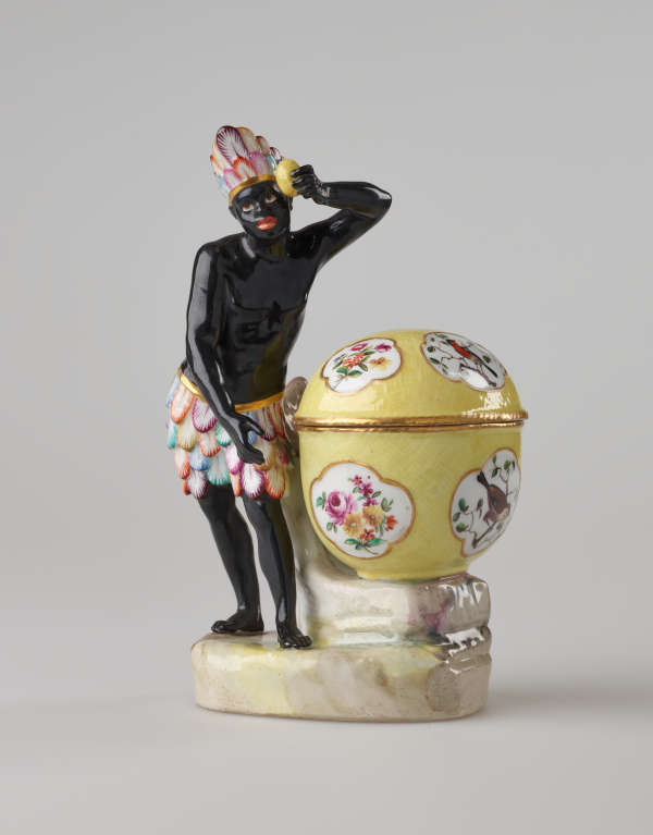 A sculptural figure with black skin, a colorful headdress and skirt. This figure is standing next to a bright yellow covered sugar basket with floral decorations.