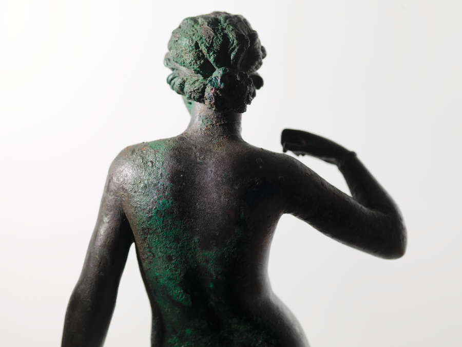 Back view of a tarnished bronze statue of a nude woman with her arm curled towards her shoulder. The lighting amplifies the texture of the statue’s surface.