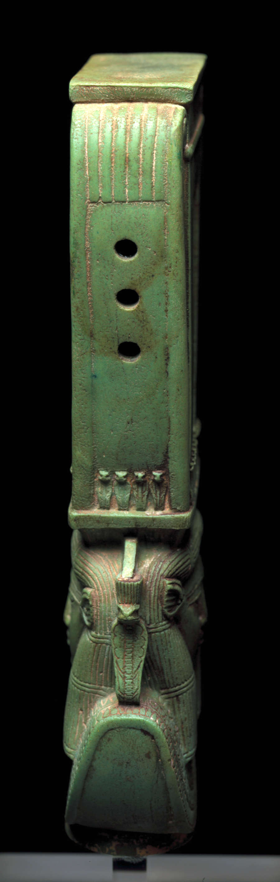 Side-view of a long jade-green sculpture depicting a collared figure’s head wearing a tall headdress. The headdress has three holds in the center and small snake figurines at the base. 