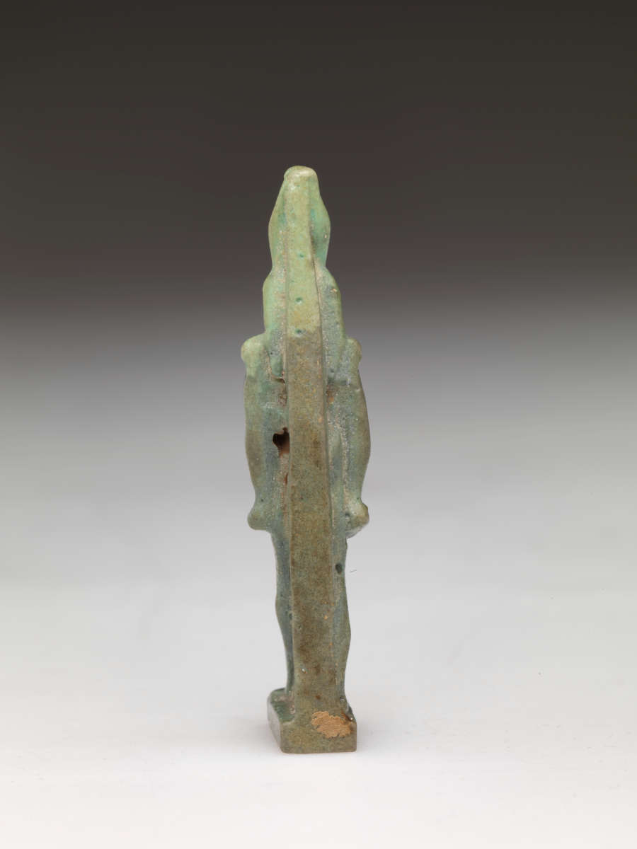 Back view of a light-jade sculpture of a standing person with a tall head, narrow torso, and straight arms. The figure is supported by a long thin sculpted rectangle extending from the base to its head.