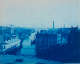 Cyanotype image of a river canal scene filled with boats, bordered by old buildings. Several industrial smokestacks emitting plumes of smoke can be seen in the distance.
