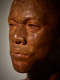 Close-up three quarter view of a detailed, wood-carved face in high-contrast lighting depicting a male figure with afrocentric facial features. The sculpture features nuanced textures, and a stern expression.