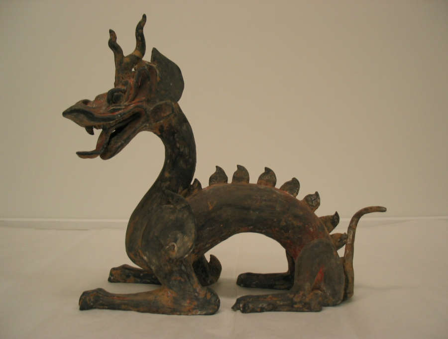 Side-view of an arched four-legged dragon with a lean body, open mouth, horned head, scaly back and a thin tail. The paint has worn into patches of browns and reds.