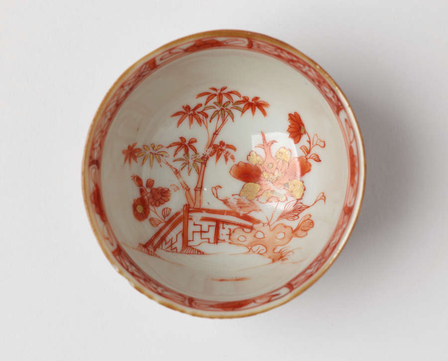 The interior of a small ceramic bowl with detailed red and gold designs with flowers, a tree, and a decorative fence, bordered by a decorative red and gold edge.