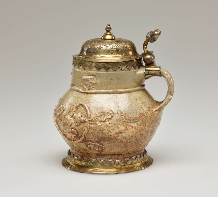 A tan colored mug with floral decoration in relief. Top is covered by a gold hinged lid and the base is surrounded by a thin gold band.