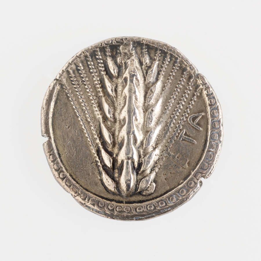 Round silver coin with irregular circle-patterned edges, embossed with an image of a grain head of wheat with splayed awns, and lettering on one side.