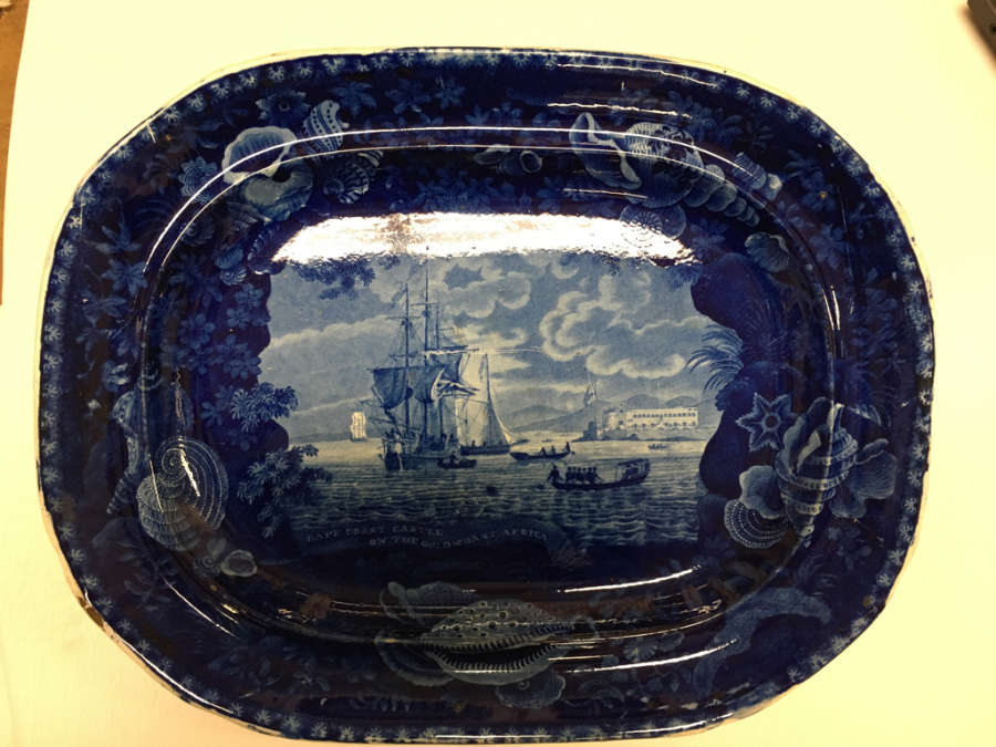 A oval shaped ceramic platter with dark blue glaze and detailed illustrations of ships and boats on water, framed by seashells and floral motifs.