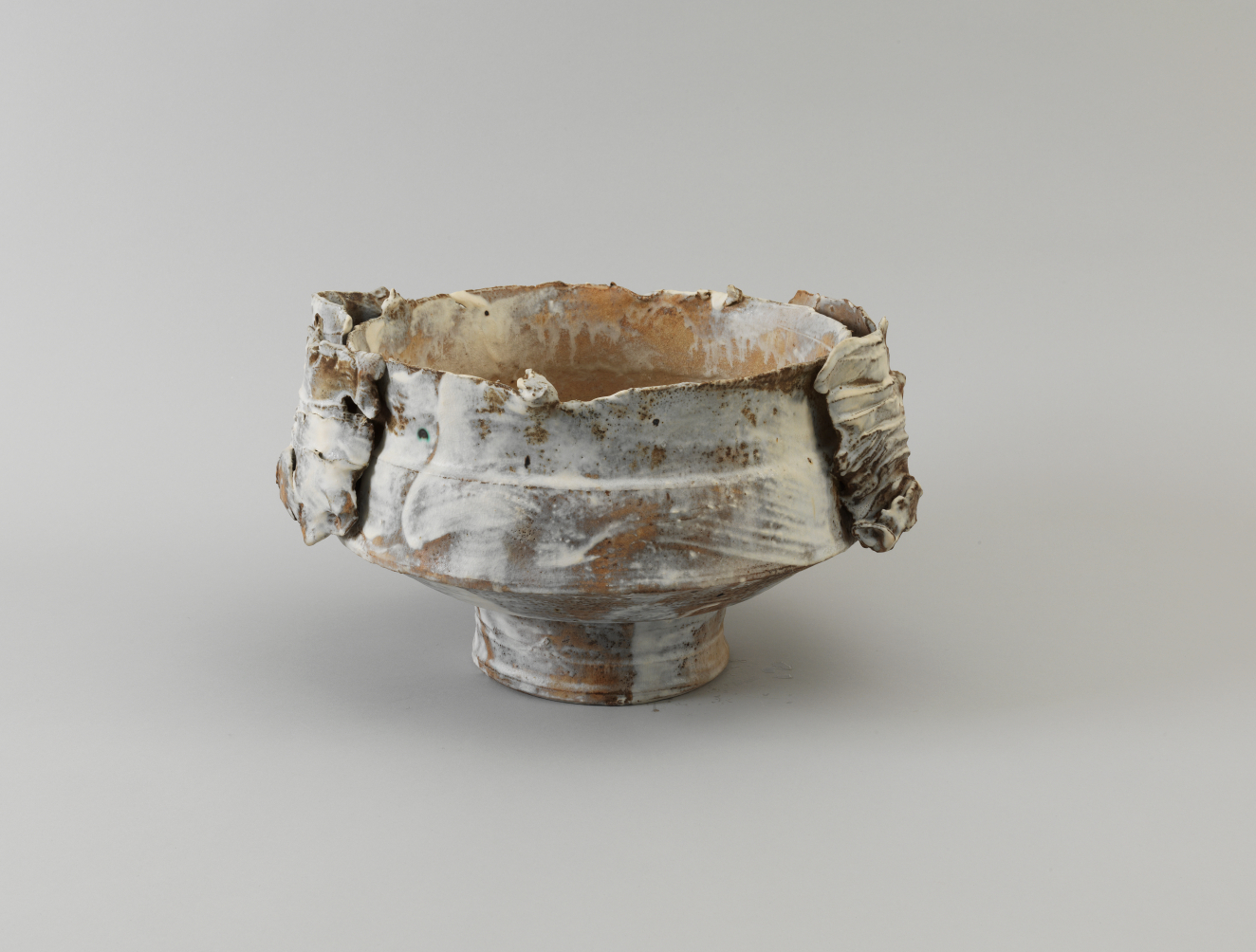 Ceramic bowl with a short pedestal base, jagged rim, and draped folds along the sides. The surface is glazed with muted gold and dry-brushed patches of white.