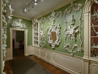 The green walls of a stately interior space featuring cream-colored woodwork are elaborately decorated with mirrors and delicate white three-dimensional figural embellishments. A built-in glass-doored cabinet is at right. 