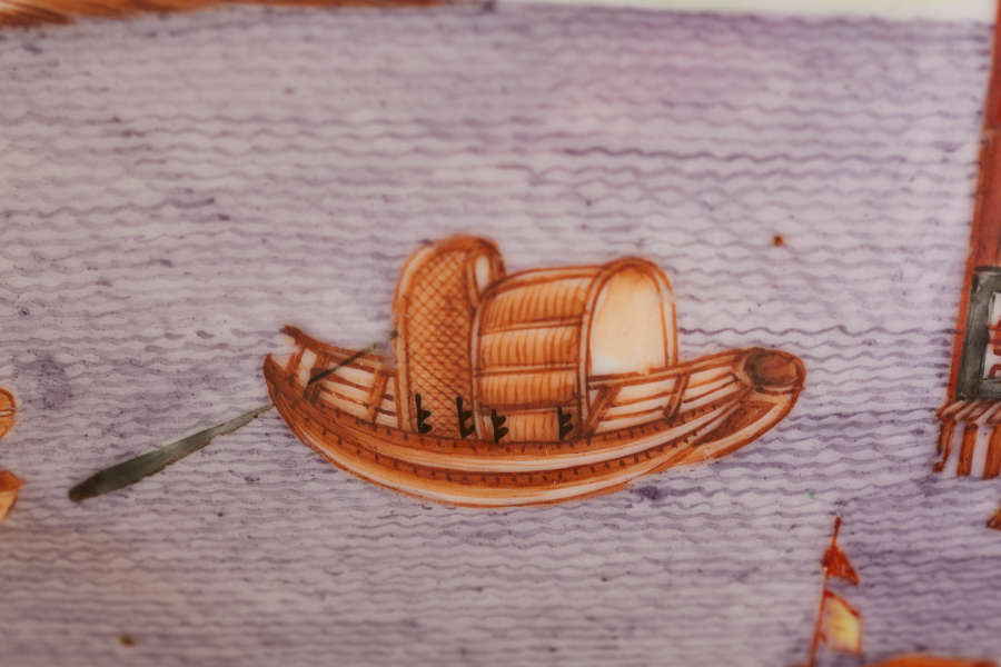 A close up of a painted ceramic depicting a small wooden boat with two arched woven canopies, floating on purple rippled water with an oar extending off the side.