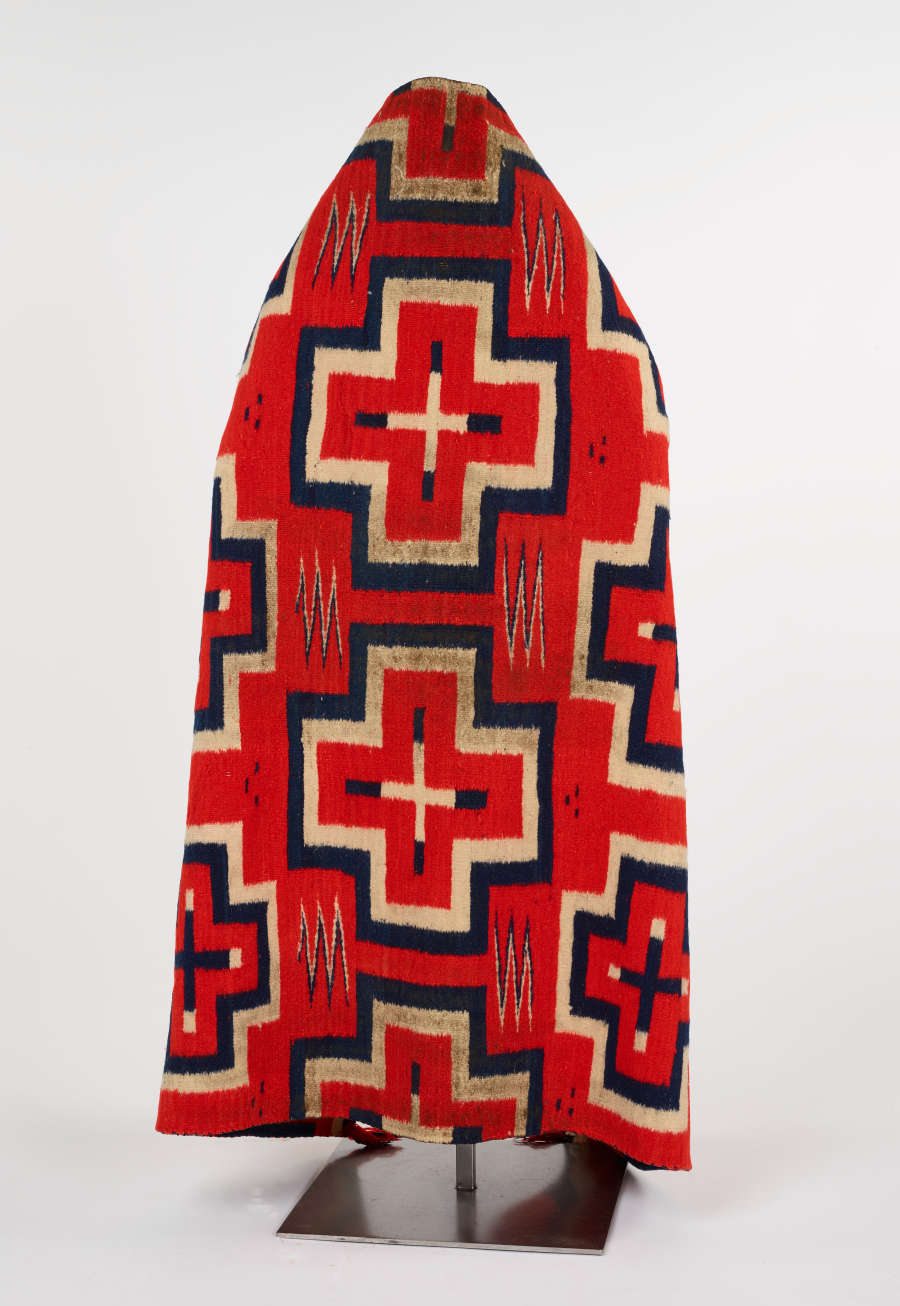 Photo of a red, patterned, woven blanket, with navy blue and cream bordered cross patterns lining the back of a mannequin. Zigzags and a three dot pattern surround the crosses.