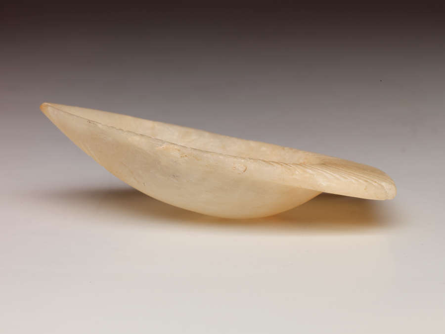 Side view of the tan bowl, showing that part of the bowl shaped as the tail of a fish extends further outwards than the rest of the bowl. It is slightly tilted.
