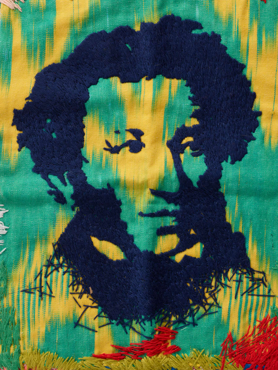 Vivid rectangular design. A man’s portrait is printed in black at center over greens and yellows. Surrounding markings in many colors, with an embroidered tree at left. Pompom-trim border.
