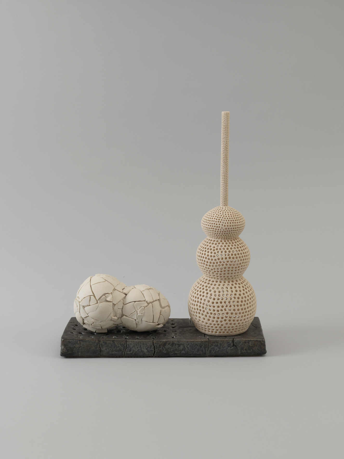 Two joined pale spheres with a cracked surface lie beside a vertical rod supporting a stack of three smooth, cream-colored spheres, all arranged on a black rectangular base.