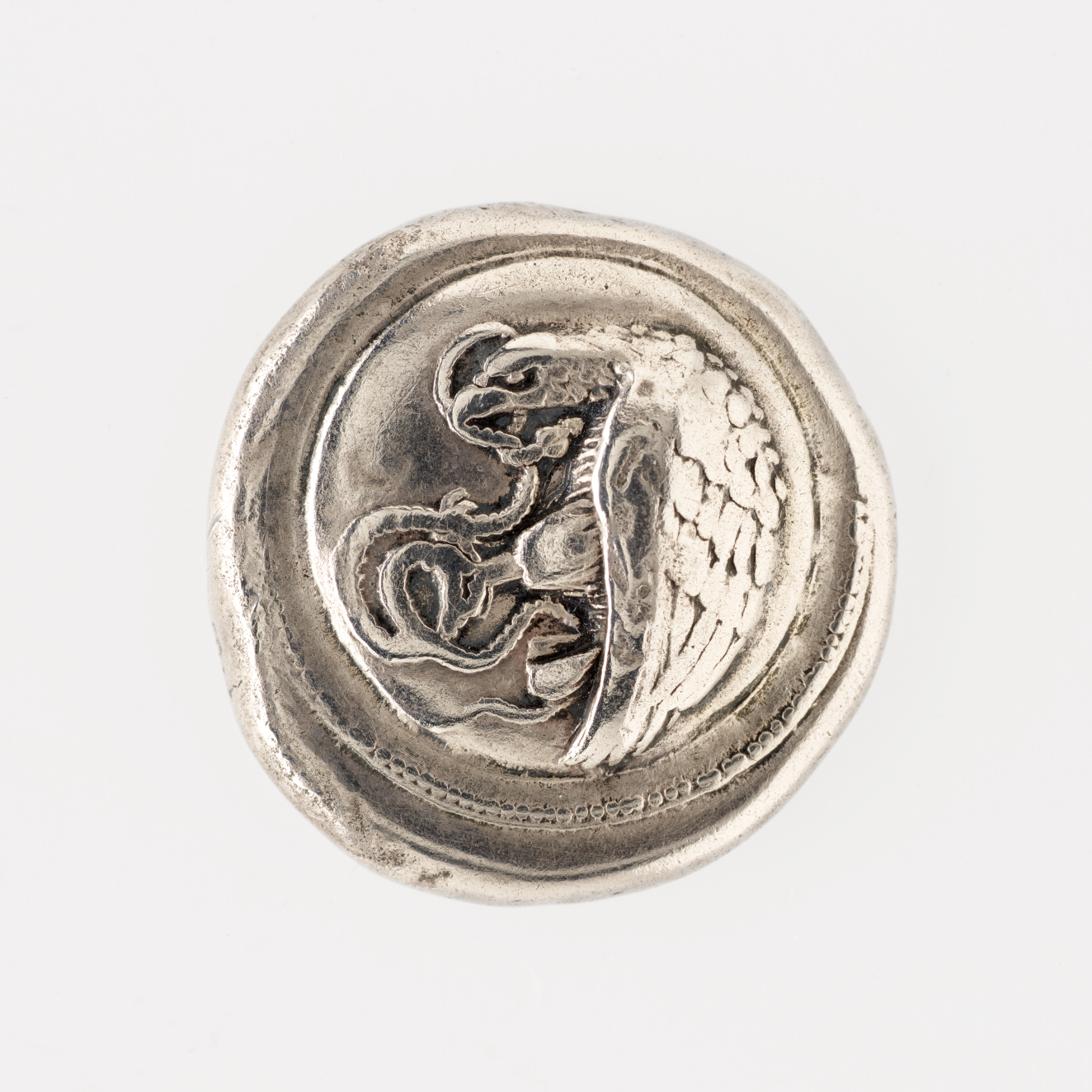 Irregular round silver coin embossed with a depiction of an eagle, curved against a circular depressed border, with a snake in its hands and mouth.