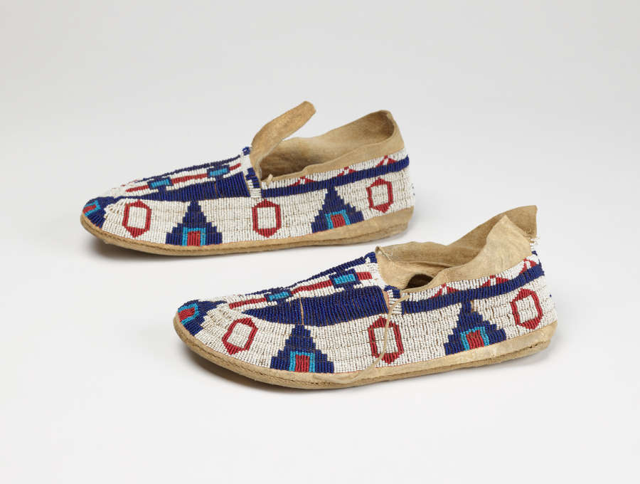 Pair of fabric shoes side-by-side and in profile, made from beaded fabric with geometric patterns in red, deep and light blues. The moccasins’ mouths are open and have two laces.