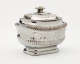 A silver sugar bowl with a decorative lid and a rounded square body with sculptural decorations.